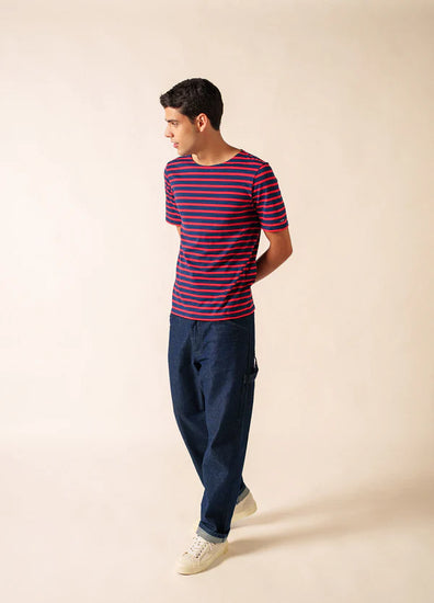Ein Mann trägt die SAINT JAMES Levant marinière mit blauer Hose und weißen Turnschuhen vor einem beigen Hintergrund.