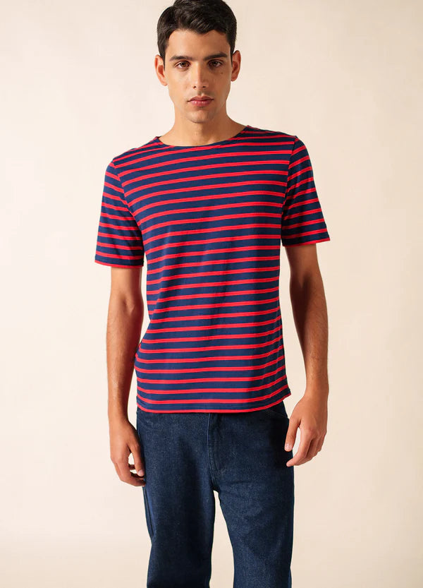 Mann in SAINT JAMES Levant marineblauem und rotem T-Shirt mit blauer Hose, stehend vor einfarbig beigem Hintergrund.