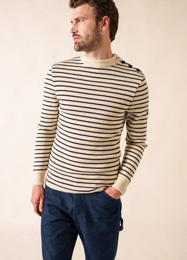 Mann in einem cremefarbenen und marineblauen SAINT JAMES Matelot Seemannspullover mit Schulterknöpfen, blaue Hose, Hand in der Tasche.