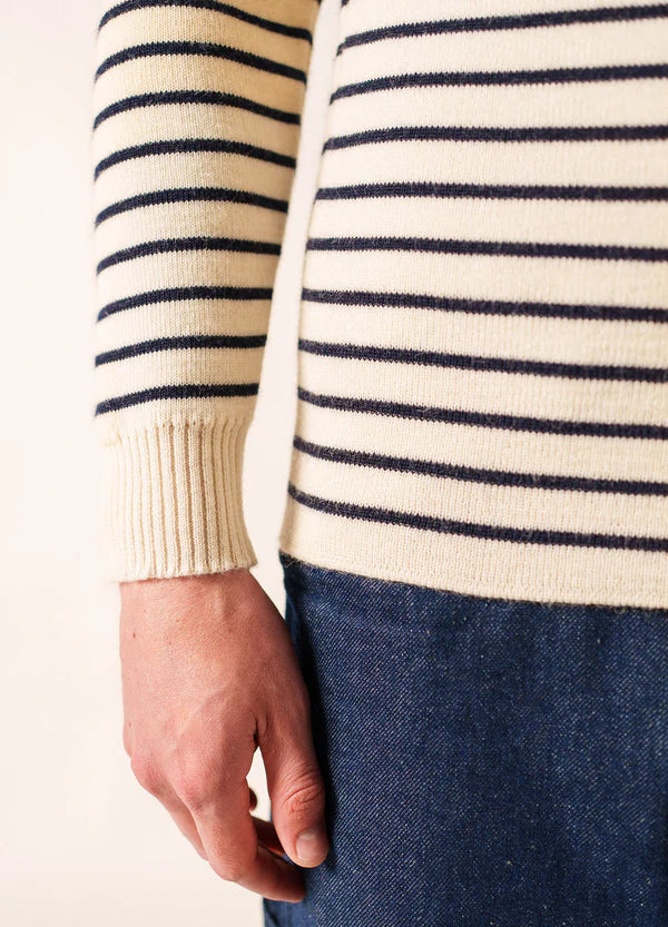 Eine Person in einem cremefarbenen SAINT JAMES Matelot mit marineblauen Streifen und blauen Jeans zeigt ihren linken Arm und ihre Hand.