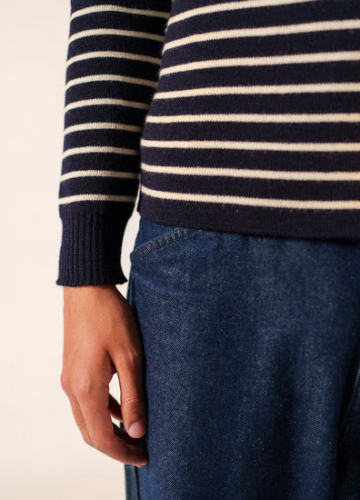 Sie trägt einen marineblauen und weißen Pullover von SAINT JAMES Matelot und eine blaue Jeans, die linke Hand ruht an ihrer Seite.