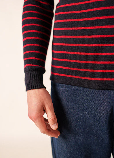 Person trägt ein schwarzes SAINT JAMES Matelot mit roten Streifen und blaue Jeans, Brust bis Mitte Oberschenkel, eine Hand sichtbar.