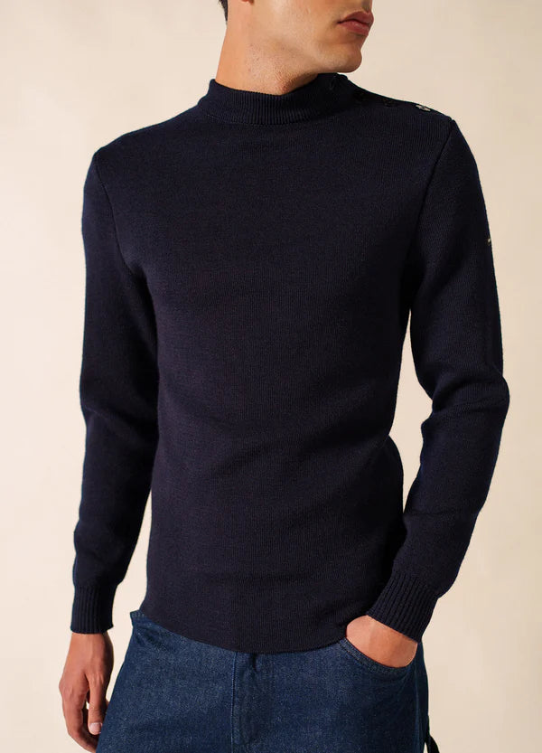 Mann trägt den marineblauen SAINT JAMES Matelot-Pullover mit Knopfleiste und blaue Jeans vor einem beigen Hintergrund.