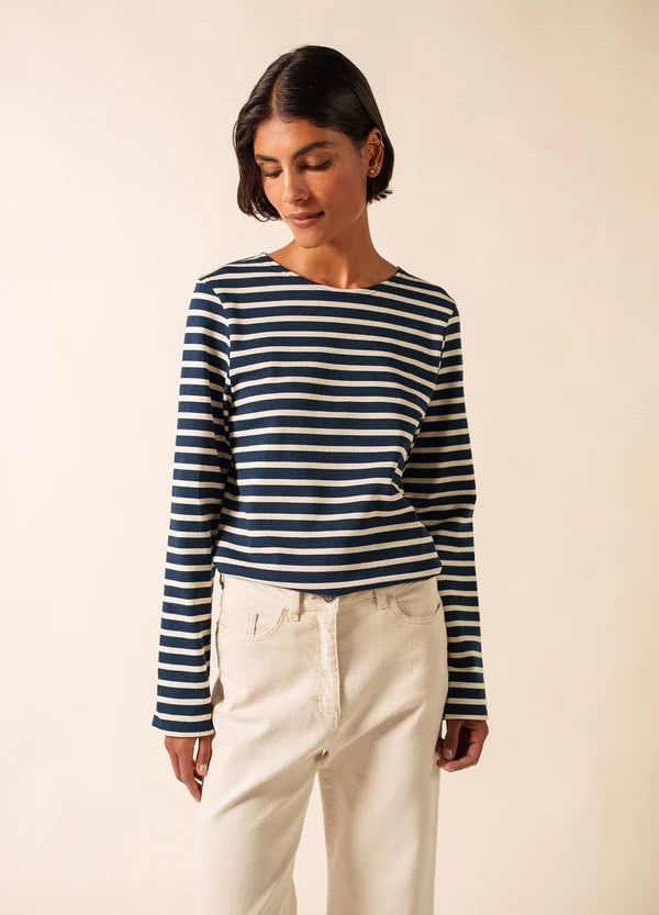 Frau in SAINT JAMES Meridien Moderne marineblaues und weißes Marinière-Hemd mit cremefarbener Hose vor einfarbigem Hintergrund.