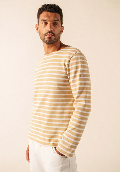 Mann, der ein beige-weiß gestreiftes langärmeliges SAINT JAMES Meridien Moderne Hemd und eine weiße Hose trägt und zur Seite schaut.