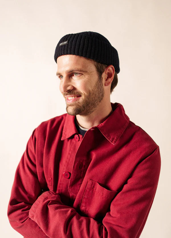 Mann in roter Jacke und schwarzer SAINT JAMES Miki Maille Mütze, lächelnd, mit Blick zur Seite vor einfarbigem Hintergrund.