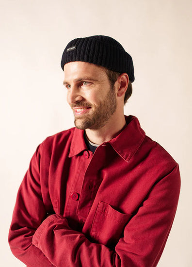 Mann in roter Jacke und schwarzer SAINT JAMES Miki Maille Mütze, lächelnd, mit Blick zur Seite vor einfarbigem Hintergrund.