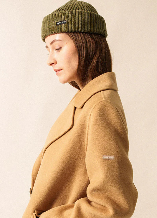 Frau in hellbraunem Mantel und SAINT JAMES Miki Maille Mütze blickt vor einem schlichten, hellen Hintergrund nach unten.