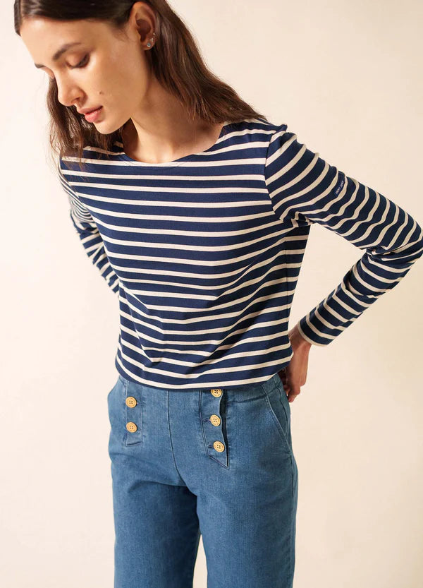 Frau in einem marineblauen SAINT JAMES Minquidame marinière und einer blauen Hose mit goldenen Knöpfen, stehend vor einem schlichten Hintergrund.