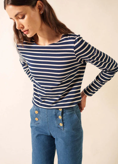 Frau in einem marineblauen SAINT JAMES Minquidame marinière und einer blauen Hose mit goldenen Knöpfen, stehend vor einem schlichten Hintergrund.