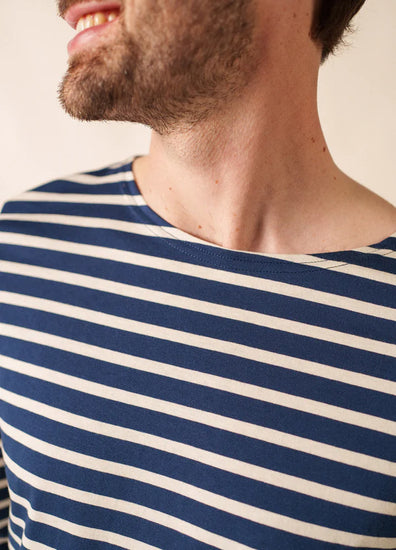 Lächelnder Mann in einem blau-weißen SAINT JAMES Minquiers-Hemd, Unisex-Modell, mit hochgezogenem Hals und teilweise sichtbarem Gesicht.