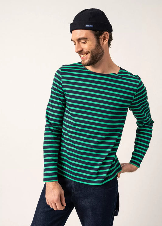 Lächelnder Mann mit schwarzer Mütze und grün gestreiftem SAINT JAMES Minquiers-Hemd, eine Hand in die Hüfte gestützt.