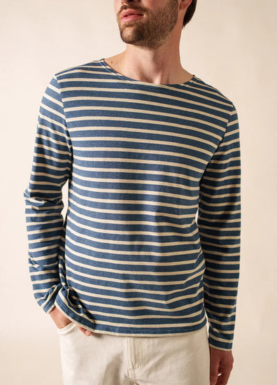 Mann mit blau-weißem SAINT JAMES Minquiers-Hemd und heller Hose, eine Hand in der Tasche.