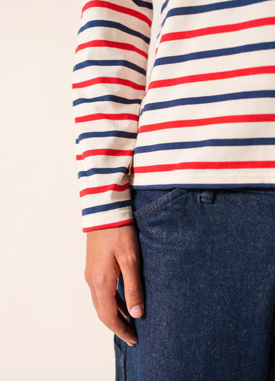 Eine Person in einem gestreiften langärmeligen SAINT JAMES Minquier Tri Unisex-Hemd und blauen Jeans, dargestellt von der Schulter bis zur Mitte des Oberschenkels.