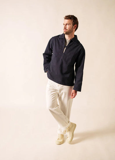 Ein Mann steht vor einem schlichten, hellen Hintergrund. Er trägt den marineblauen SAINT JAMES Nemo-Pullover mit kurzem Reißverschluss, dazu eine weiße Leinenhose und cremefarbene Turnschuhe. Er schaut nach links, eine Hand in der Tasche und die Beine lässig übereinander geschlagen.