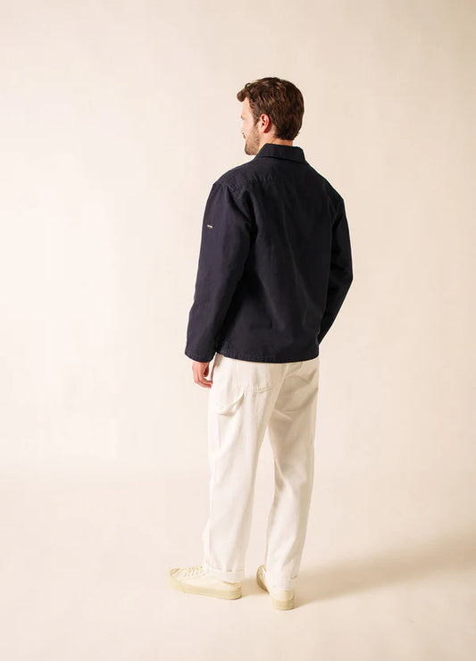 Ein Mann blickt in die Ferne. Er trägt die dunkle marineblaue Nemo-Jacke von SAINT JAMES in maritimer Tradition, gepaart mit einer weißen Hose und cremefarbenen Schuhen vor einem schlichten hellen Hintergrund.