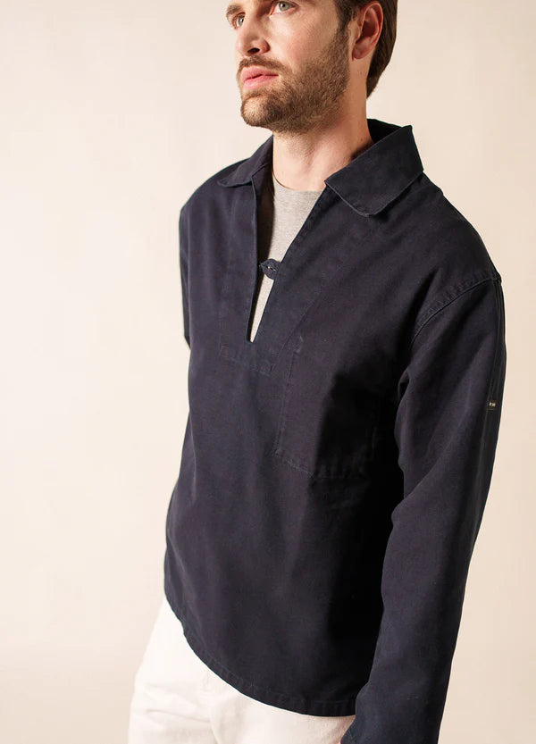 Ein Mann, der den marineblauen Segeltuchpullover SAINT JAMES Nemo über einem grauen Oberteil und einer weißen Hose trägt, steht vor einem beigen Hintergrund, der die maritime Tradition widerspiegelt, während sein Blick leicht nach oben gerichtet ist.