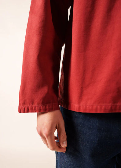 Nahaufnahme einer Person, die den roten, langärmeligen SAINT JAMES Nemo Fischerkittel und dunkelblaue Jeans trägt, die linke Hand entspannt an der Seite vor einem schlichten, hellen Hintergrund, der an maritime Traditionen erinnert.