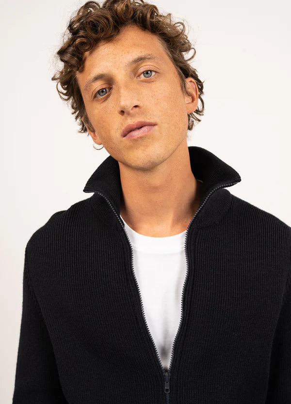 Junger Mann mit lockigem Haar trägt die schwarze Wollstrickjacke Quiberon von SAINT JAMES über weißem T-Shirt.