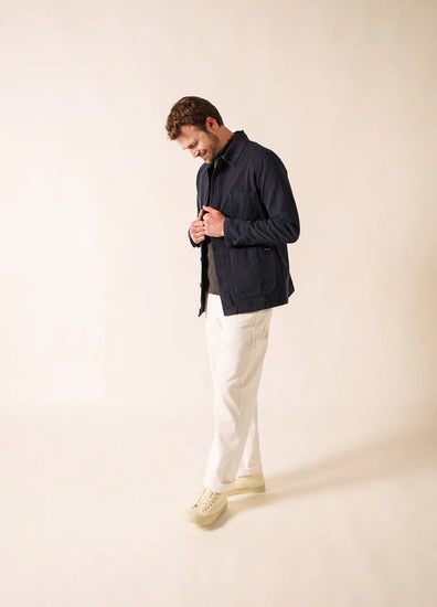 Mann im Profil, nach unten blickend, trägt die SAINT JAMES Sirocco Jacke, weiße Hosen und cremefarbene Turnschuhe vor einem hellen Hintergrund.