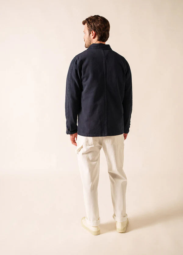 Mann in marineblauer SAINT JAMES Sirocco-Jacke, weißer Hose und Schuhen vor einem schlichten hellen Hintergrund.