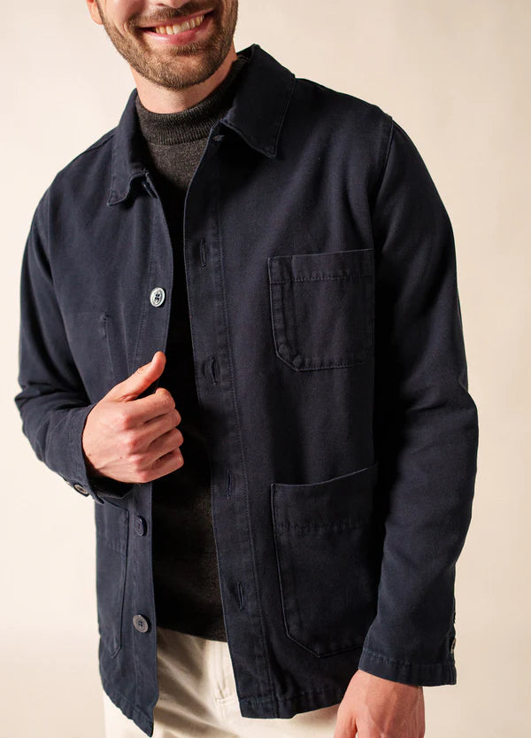 Mann lächelnd in SAINT JAMES Sirocco navy blue Canvas Jacke über einem dunklen Pullover und weißer Hose, vor einfarbigem Hintergrund.