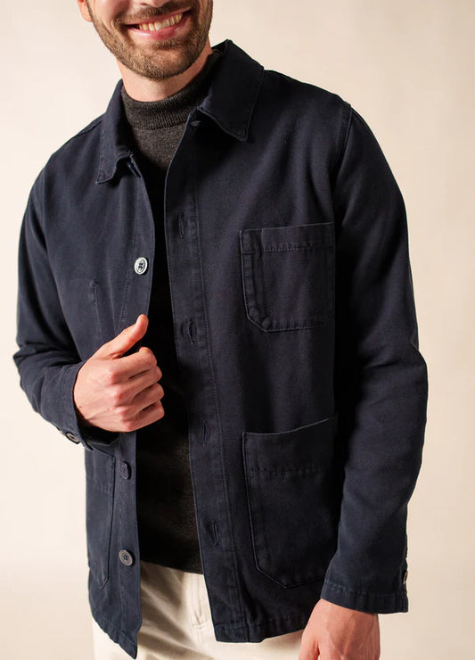 Mann lächelnd in SAINT JAMES Sirocco navy blue Canvas Jacke über einem dunklen Pullover und weißer Hose, vor einfarbigem Hintergrund.