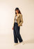 Eine Frau im gestreiften SAINT JAMES St. Cathy-Shirt, einer Leinenjacke, einer marineblauen Hose und Turnschuhen steht vor einem schlichten Hintergrund.
