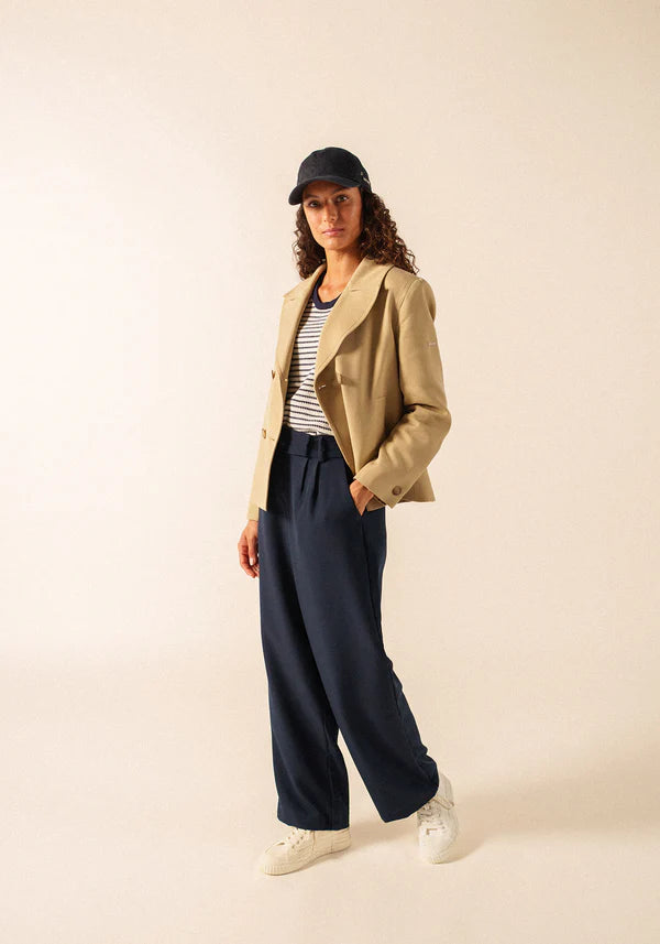 Eine Frau im gestreiften SAINT JAMES St. Cathy-Shirt, einer Leinenjacke, einer marineblauen Hose und Turnschuhen steht vor einem schlichten Hintergrund.