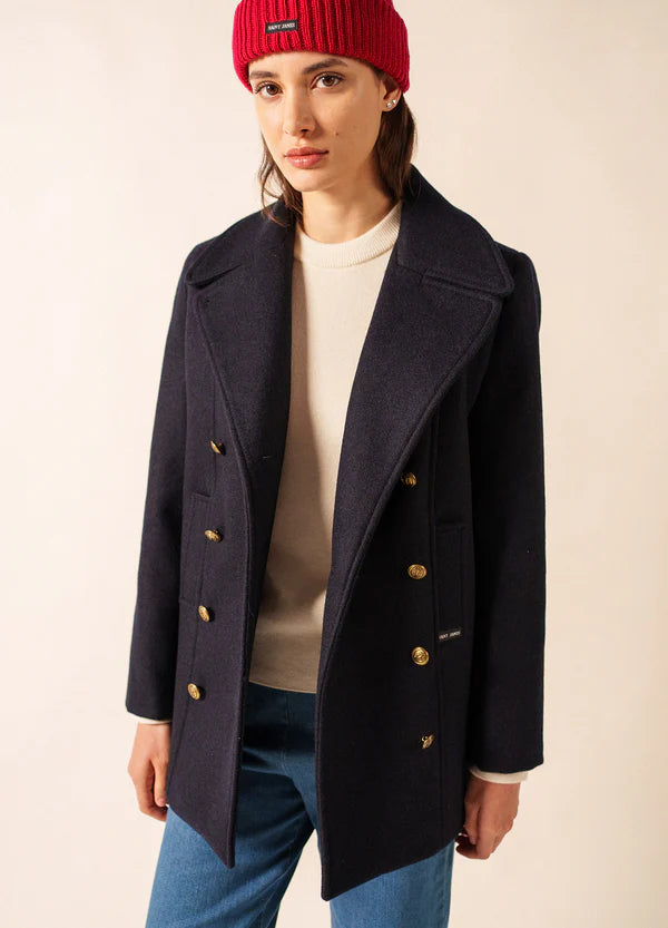 Frau in einem marineblauen Wollmantel von SAINT JAMES Voilure, roter Mütze und blauen Jeans vor einem hellen Hintergrund.