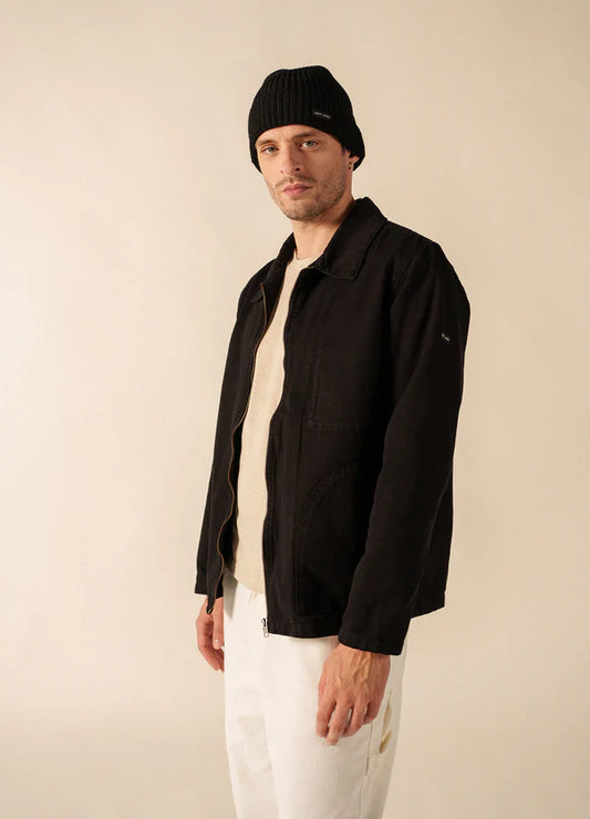 Ein Mann in einer schwarzen SAINT JAMES Zephyr-Jacke, einem beigen Hemd, einer weißen Hose und einer schwarzen Mütze steht vor einem schlichten Hintergrund.