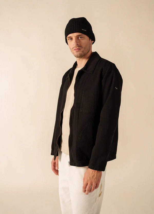 Ein Mann in einer schwarzen SAINT JAMES Zephyr-Jacke, einem beigen Hemd, einer weißen Hose und einer schwarzen Mütze steht vor einem schlichten Hintergrund.