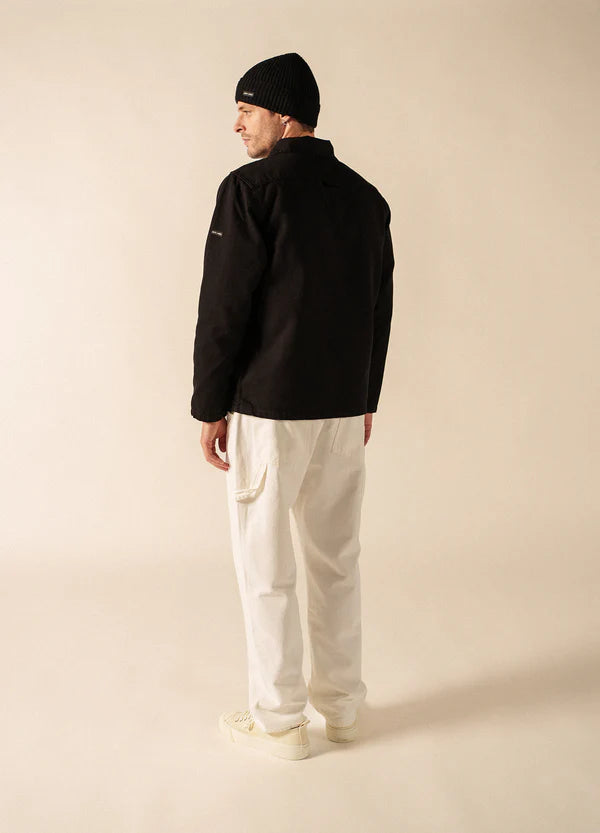 Ein Mann trägt eine schwarze SAINT JAMES Zephyr-Mütze, eine schwarze Jacke und eine weiße Hose und schaut in einer neutralen Studioumgebung weg.