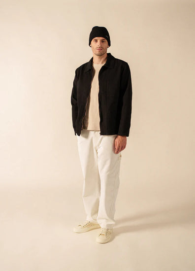 Mann im Studio trägt SAINT JAMES Zephyr, schwarze Canvasjacke, weiße Hose, beiges Hemd, Turnschuhe und schwarze Mütze.