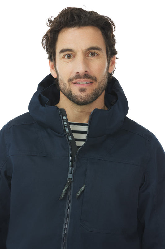 Mann mit lockigem braunem Haar trägt das Mousqueton Ardan marineblauer Parka und gestreiftes Hemd, stehend vor einem weißen Hintergrund.