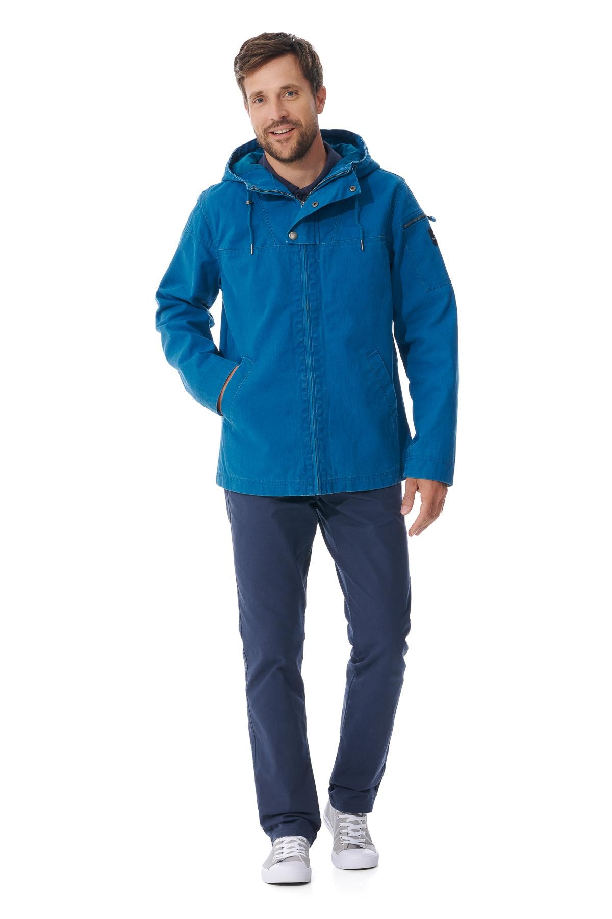 Mann in blauer wasserabweisender Jacke von MOUSQUETON Baden, marineblaue Hose, graue Turnschuhe, lächelnd mit Hand in der Tasche.