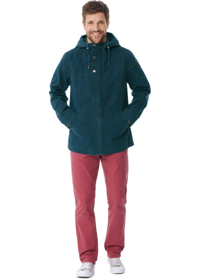 Mann in MOUSQUETON Baden, blaugrüne Jacke, rote Hose und weiße Turnschuhe, Hände in den Taschen, lächelnd.