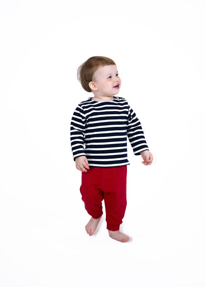 Kleinkind in gestreiftem Baumwollhemd und roter Hose von MOUSQUETON Mario Bebe, barfuß vor weißem Hintergrund.