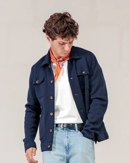 Ein Mann trägt die marineblaue Strickjacke „Lois“ von ROYAL MER, ein weißes Hemd, Jeans und ein orangefarbenes Bandana; er blickt mit der Hand in der Hosentasche nach unten.