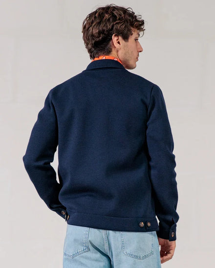 Ein Mann in einer marineblauen Merinowolle-Jacke der Marke ROYAL MER „Lois“ und hellblauen Jeans, mit dem Rücken zur Kamera, vor einem neutralen Hintergrund.