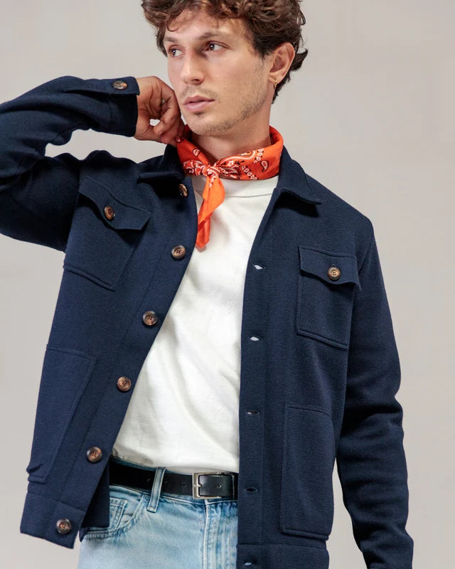 Ein Mann in einer marineblauen Strickjacke von ROYAL MER Lois, einem weißen Hemd, hellen Jeans und einem orangefarbenen Bandana blickt zur Seite, wobei seine Hand in der Nähe seines Halses liegt.