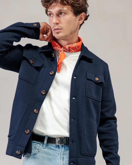 Ein Mann in einer marineblauen Strickjacke von ROYAL MER Lois, einem weißen Hemd, hellen Jeans und einem orangefarbenen Bandana blickt zur Seite, wobei seine Hand in der Nähe seines Halses liegt.