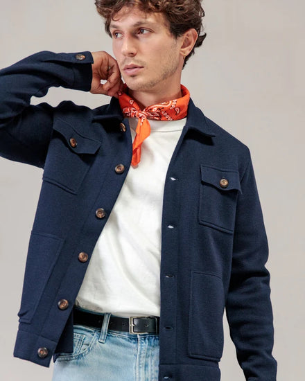 Ein Mann in einer marineblauen Strickjacke von ROYAL MER Lois, einem weißen Hemd, hellen Jeans und einem orangefarbenen Bandana blickt zur Seite, wobei seine Hand in der Nähe seines Halses liegt.