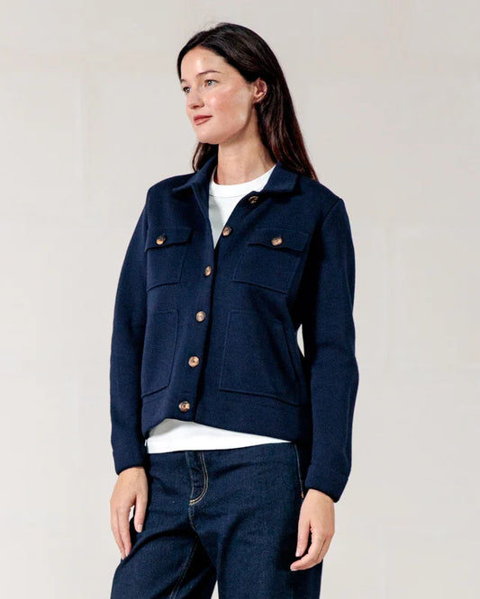 Eine Frau in der Merinowoll-Strickjacke „Noyale“ von ROYAL MER trägt Jeans vor einem hellen, minimalistischen Hintergrund.