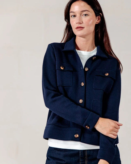 Eine Frau in einer marineblauen Noyale-Jacke von ROYAL MER über einem weißen Oberteil, die vor einem schlichten, hellen Hintergrund steht.
