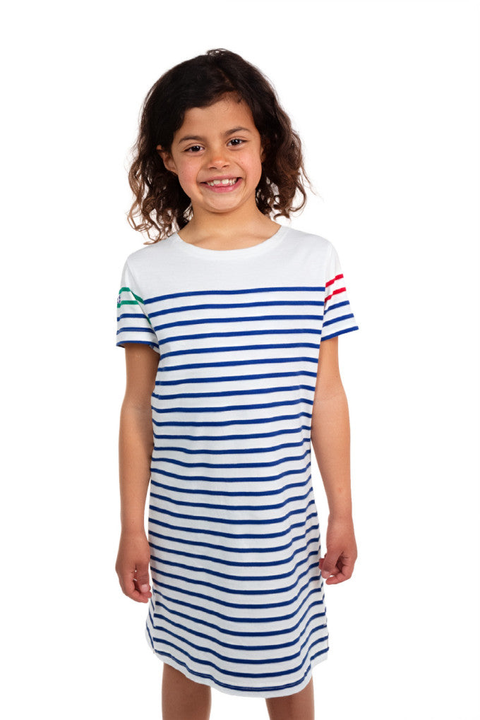 Lächelndes Kind in einem mousqueton Tymarianig Kleid mit blauen Streifen, stehend vor einem weißen Hintergrund.
